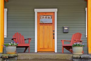 A home entryway features a porch with two red chairs on each side of a wood entry door. The home features green vinyl siding.
